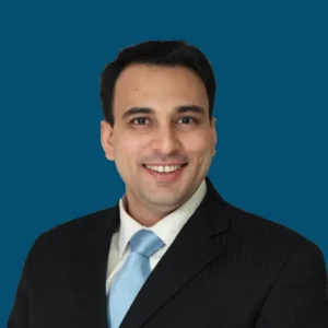 Smiling man in a black suit with a light blue tie against a solid blue background