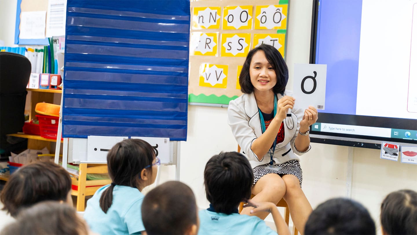 A teacher sitting and engaging with young students in a classroom, holding up a card with a letter while teaching phonics, with a colorful educational display in the background.