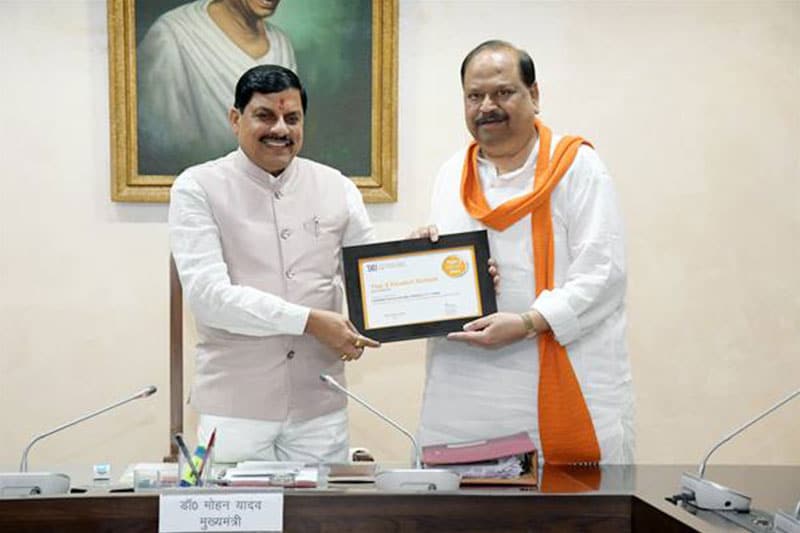 Two officials smiling and holding a framed certificate during a formal event in an office setting.