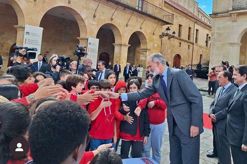 King of Spain greeting children in red shirts during a public event at a historic courtyard, surrounded by officials and media.