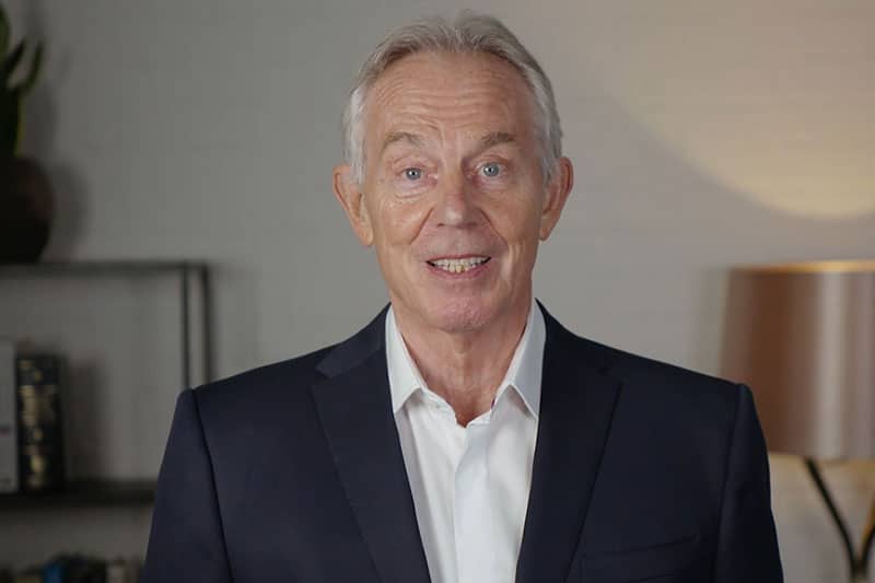Sir Tony Blair wearing a dark suit and white shirt, smiling while speaking indoors.