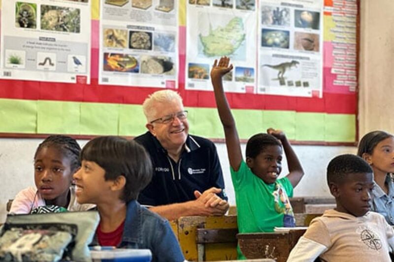 The Premier of the Western Cape, Alan Winde, sitting among students in a classroom, smiling as a student raises their hand.