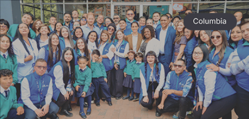 Group photo of school staff, students, officials, and Prince Harry and Meghan, Duke and Duchess of Sussex, gathered outside a building, smiling and celebrating together.