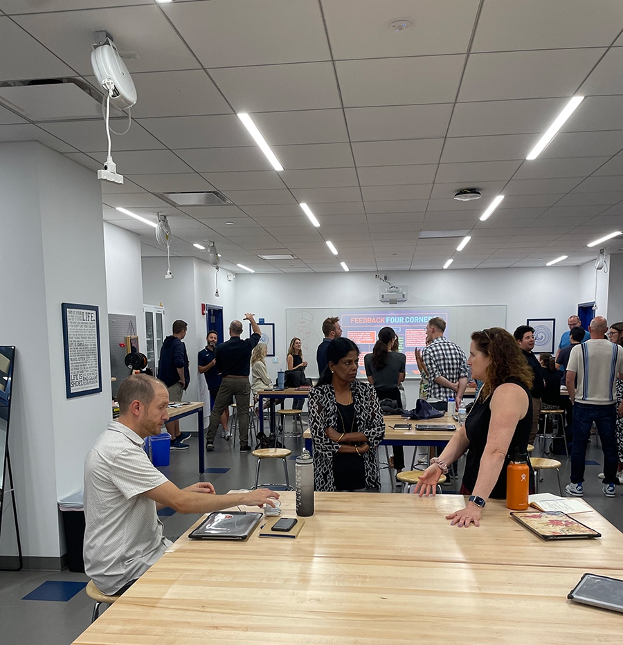 Group of people engaged in discussions and collaborative activities in a bright and modern workshop or training room, with wooden tables, tools, and a presentation displayed on a screen in the background.