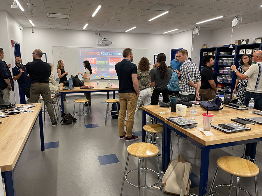 Workshop participants interacting and networking in a collaborative space with wooden tables, stools, and a presentation screen in the background displaying 'Feedback Four Corners'.