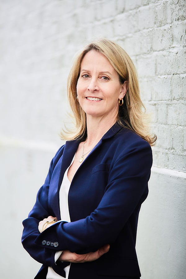 Professional portrait of Jenny Anderson, a confident woman in a navy blazer standing against a brick wall with arms crossed and a friendly smile.