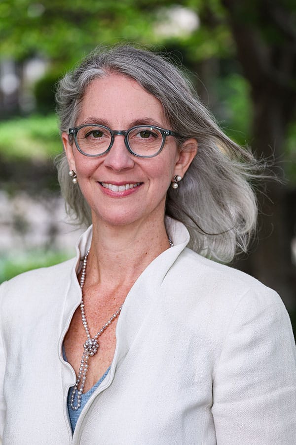 Professional portrait of Rebecca Winthrop, wearing a white blazer and standing outdoors with a blurred green background.