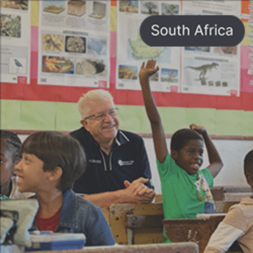 The Premier of the Western Cape, Alan Winde, sitting among students in a classroom, smiling as a student raises their hand.