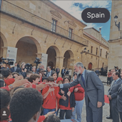 King of Spain greeting children in red shirts during a public event at a historic courtyard, surrounded by officials and media.