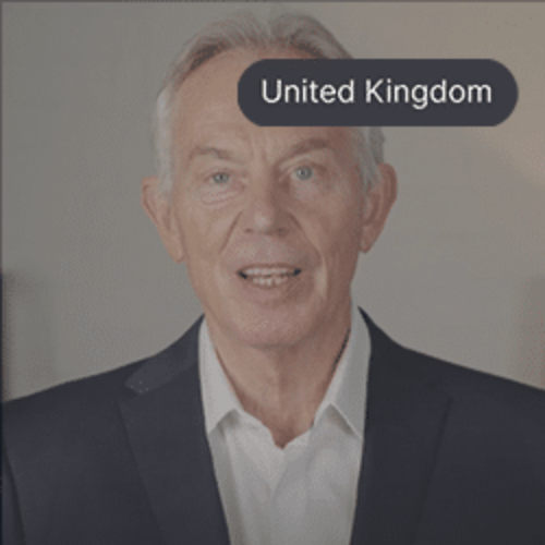 Sir Tony Blair wearing a dark suit and white shirt, smiling while speaking indoors.