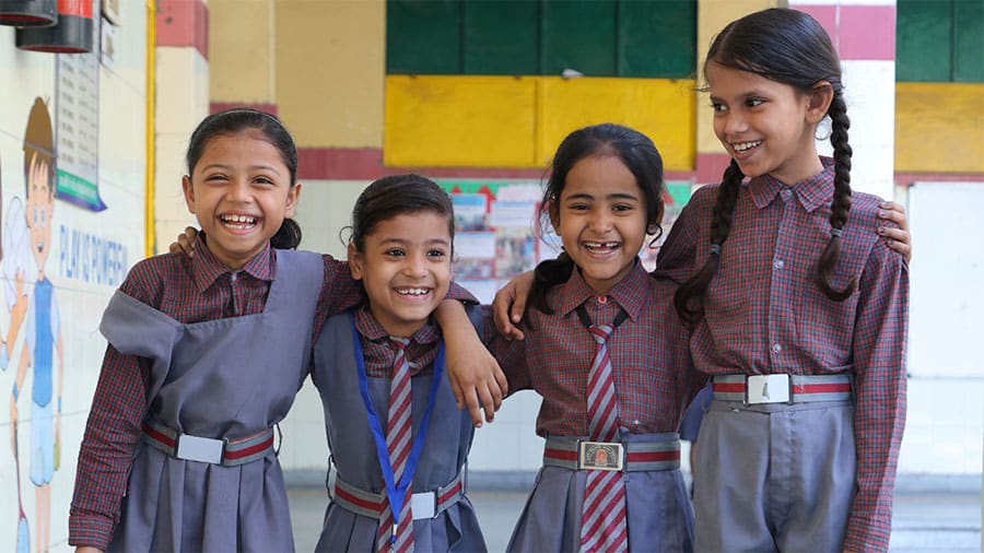 Four smiling schoolgirls in uniforms with arms around each other in a classroom setting.