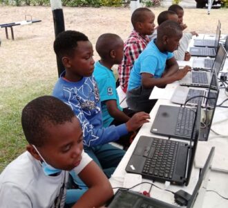 A group of young students sitting outdoors at a long table, working on laptops. They are focused on their screens, participating in a learning activity at what appears to be a technology-based educational session.