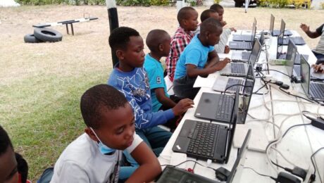 A group of young students sitting outdoors at a long table, working on laptops. They are focused on their screens, participating in a learning activity at what appears to be a technology-based educational session.