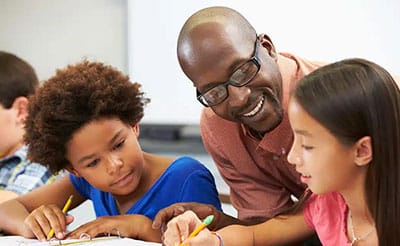 A smiling teacher wearing glasses engages with two young students, helping them with their schoolwork in a supportive and interactive learning environment.