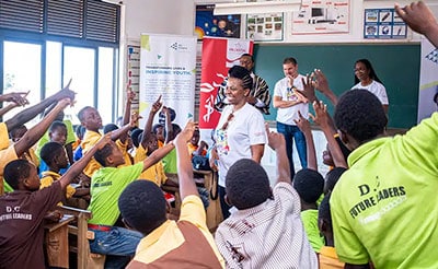 A classroom filled with enthusiastic students wearing colorful uniforms, eagerly raising their hands while engaging with facilitators during an interactive learning session.