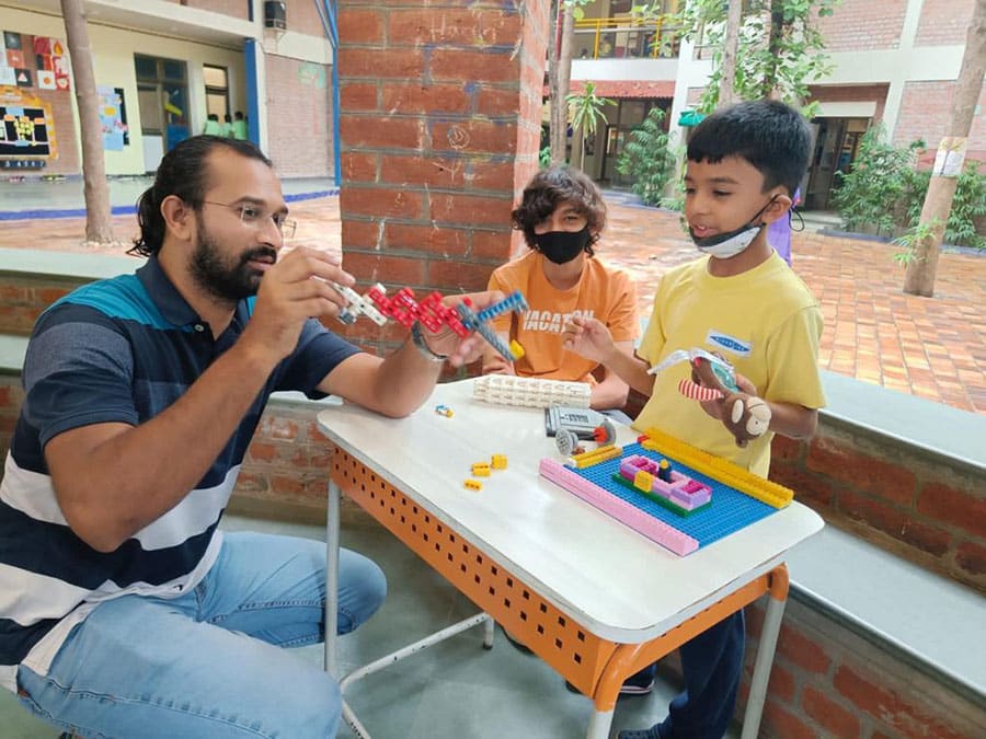 A teacher and two young students engaged in a hands-on learning activity with colorful LEGO pieces at a small table in an open, well-lit school environment. The teacher is explaining something while the students listen attentively and interact with the models.