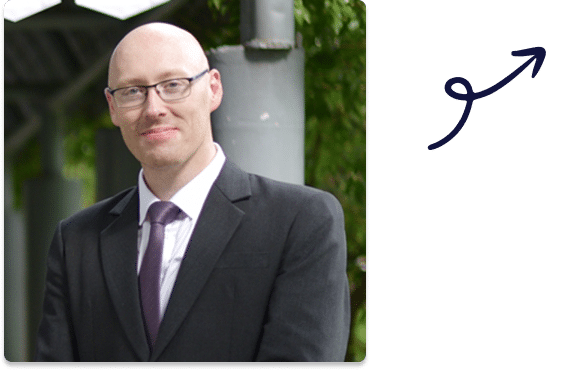 Man in a suit and tie standing outdoors, smiling confidently.