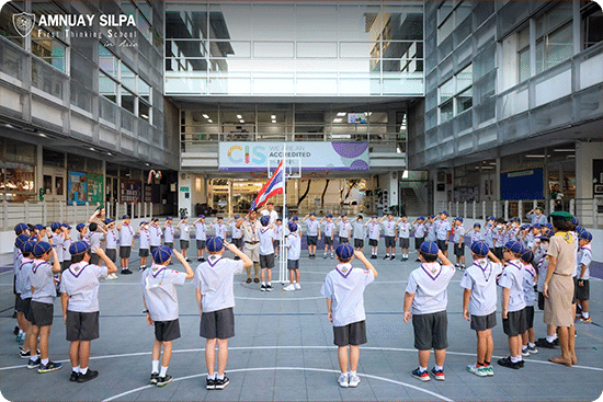 Students in scout uniforms saluting during a morning assembly at Amnuay Silpa School courtyard.