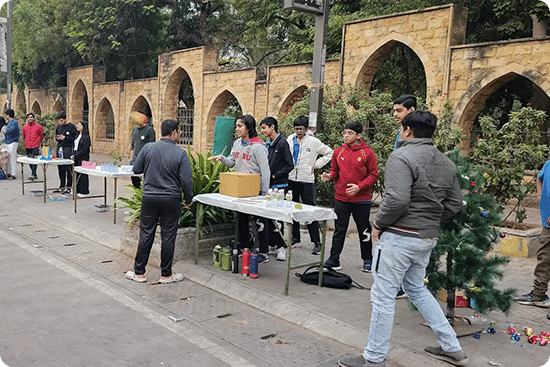 Students setting up stalls for a community event outside a school with arched stone walls in the background.