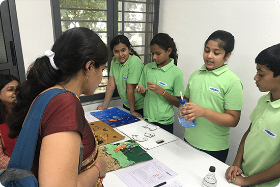 Students in green uniforms explaining a science project to a visitor at a school exhibition.