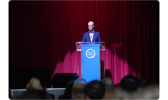 A keynote speaker delivering an address at the podium during the World Schools Summit, with a red curtain backdrop and audience in view.