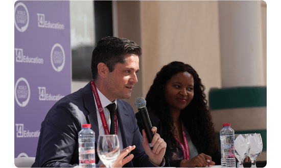 A panelist speaking into a microphone during a World Schools Summit session, with another panelist listening attentively.