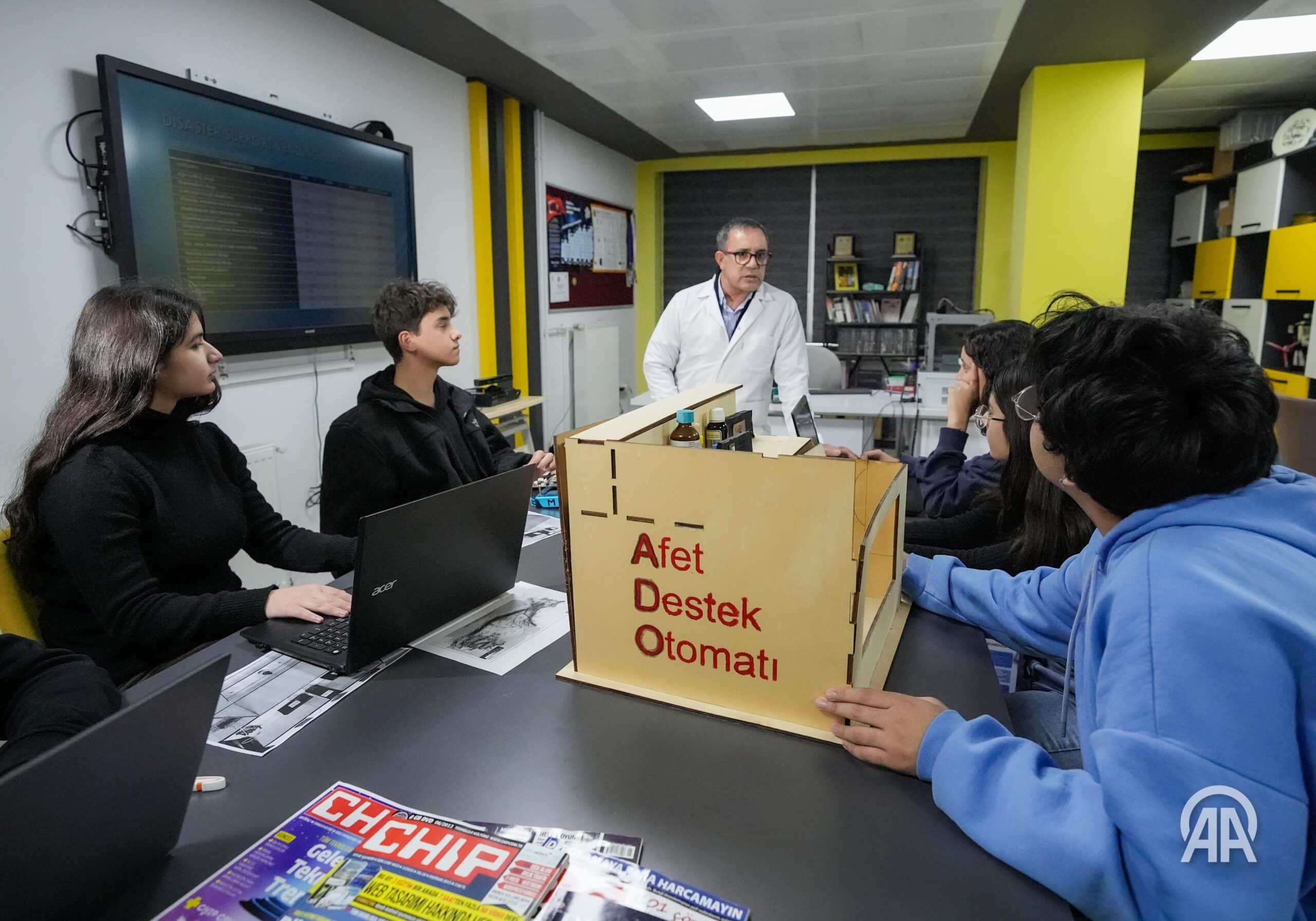 A teacher and a group of students sit around a table with laptops, discussing a project labelled 