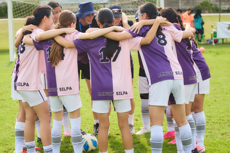 Girls' soccer team huddling together on the field before a match, wearing pink and purple jerseys