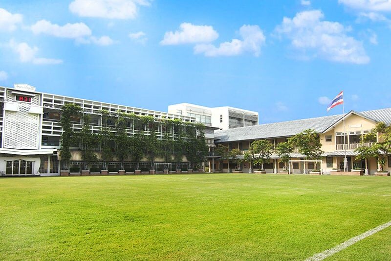 School campus with green lawn, multiple academic buildings, and a Thai flag flying under a bright blue sky