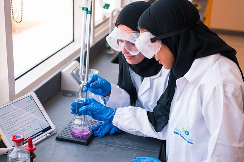Two female students in lab coats and goggles conducting a chemistry experiment at ATS UAQ