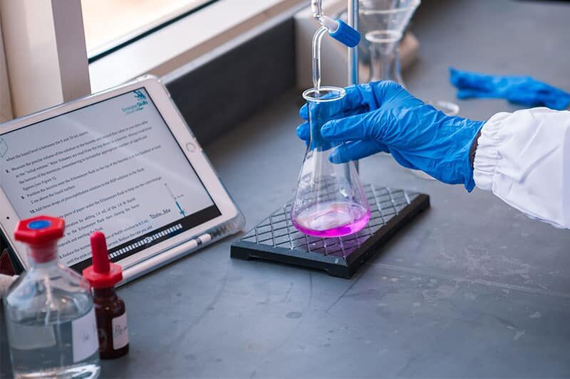 Close-up of gloved hand handling a flask during a lab experiment with digital instructions visible on tablet