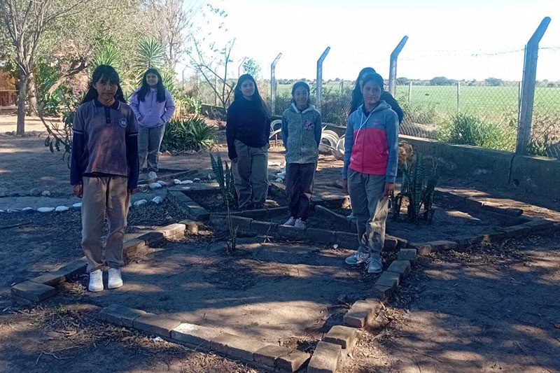 Six schoolgirls standing in a brick-outlined garden bed at a rural school garden project