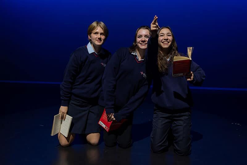 Three students performing a dramatic scene on stage with scripts in hand