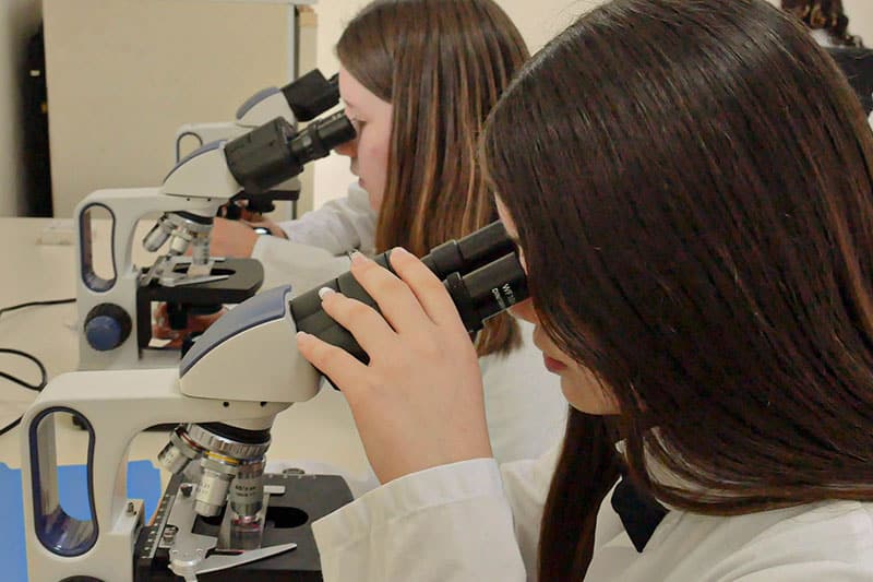 Students in lab coats using microscopes during a science class at Escuela y Colegio Científico Interamericano