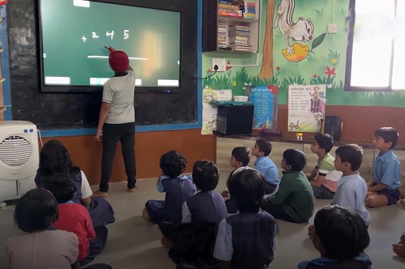 Young students sitting on the floor and watching a classmate solve a math problem on a digital board at The School Jalindarnagar