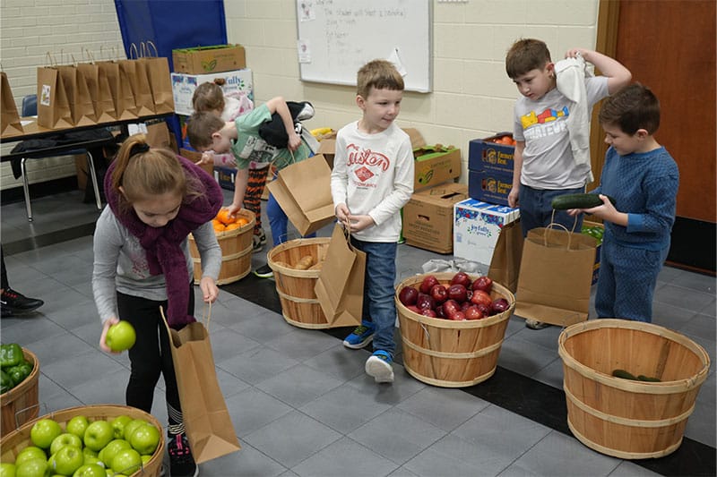 Young children collect fresh fruits and vegetables into paper bags from baskets in a classroom-like setting.