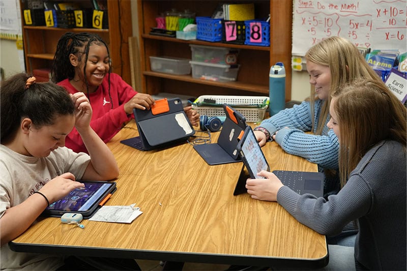Four students sitting at a classroom table using tablets for a digital learning activity, smiling and engaged in a collaborative environment.