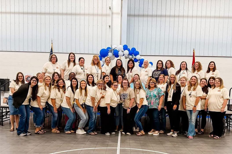Large group of school staff members posing for a group photo in a gymnasium, wearing matching shirts with a blue and white balloon arch in the background.