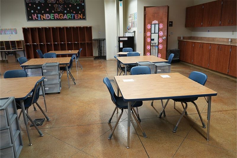 Empty kindergarten classroom with small tables and chairs arranged neatly, cubby storage along the wall, and a welcome sign above.
