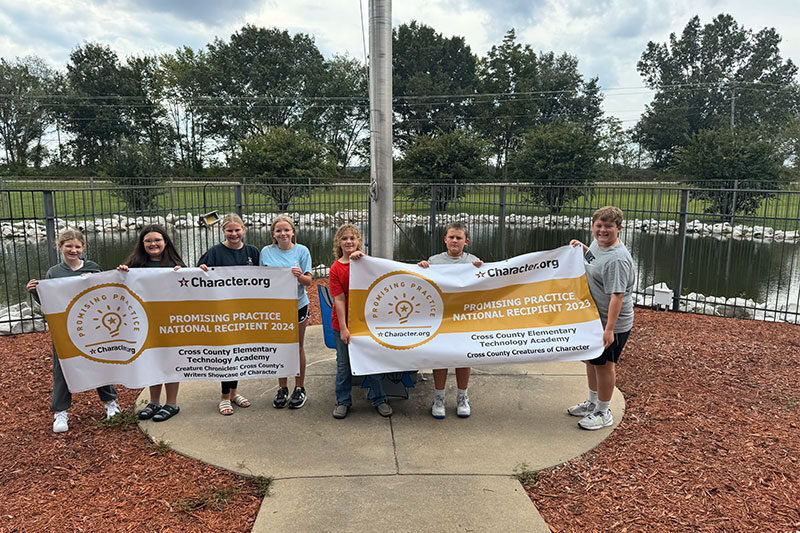 Group of students standing outdoors holding banners for Character.org's Promising Practice National Recipient awards for 2023 and 2024, in front of a pond and flagpole.