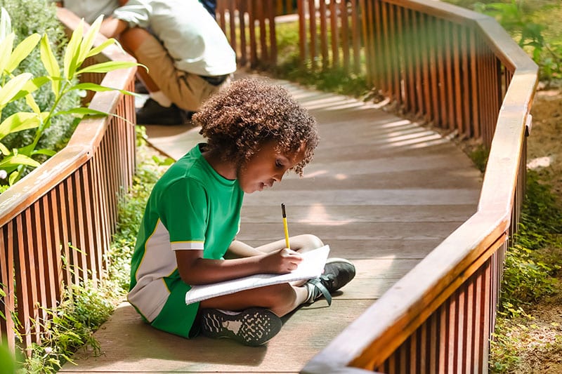 Young child sitting on a wooden path, writing in a notebook with a pencil in a garden setting.