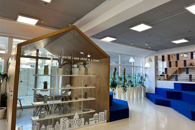 A modern, minimalist school interior with a glass-walled reading nook shaped like a house and tiered blue seating for students.