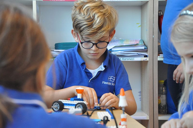 A young boy in a blue Finnish School uniform builds a small vehicle model using LEGO-style pieces in a classroom setting.
