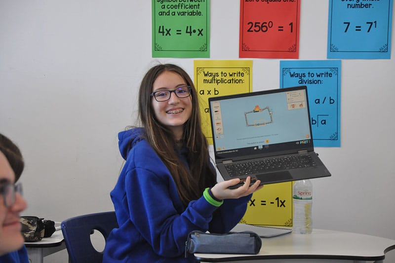 Smiling student in a blue hoodie holds up a laptop displaying an educational simulation, seated in a classroom with colourful math posters on the wall.