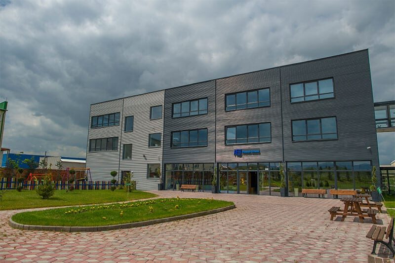 Modern three-story school building with large windows and a paved courtyard featuring benches, picnic tables, and green landscaping.