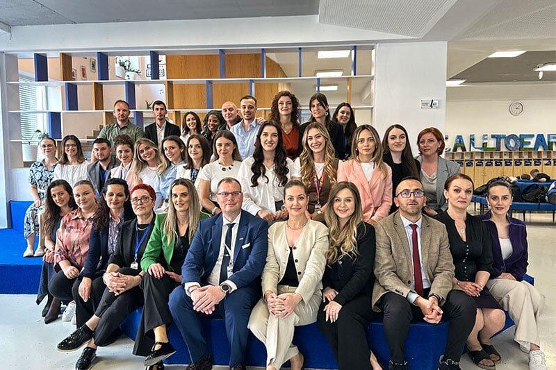 A large group of professionally dressed school staff and administrators pose together in a modern learning space with blue tiered seating and open shelving.