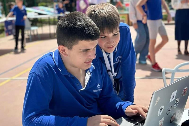 Two boys in Finnish School uniforms work together on a laptop outdoors during what appears to be a school event or project activity.