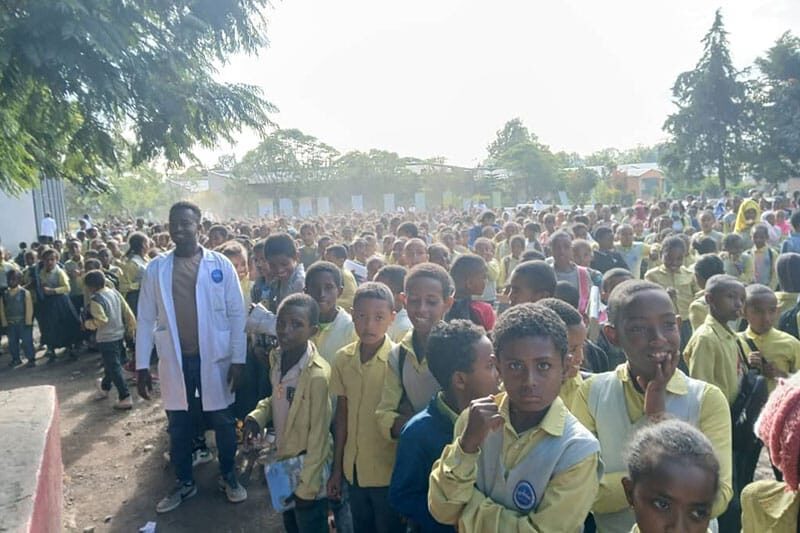 Large crowd of school children in yellow uniforms gathered outdoors with a teacher in a white coat