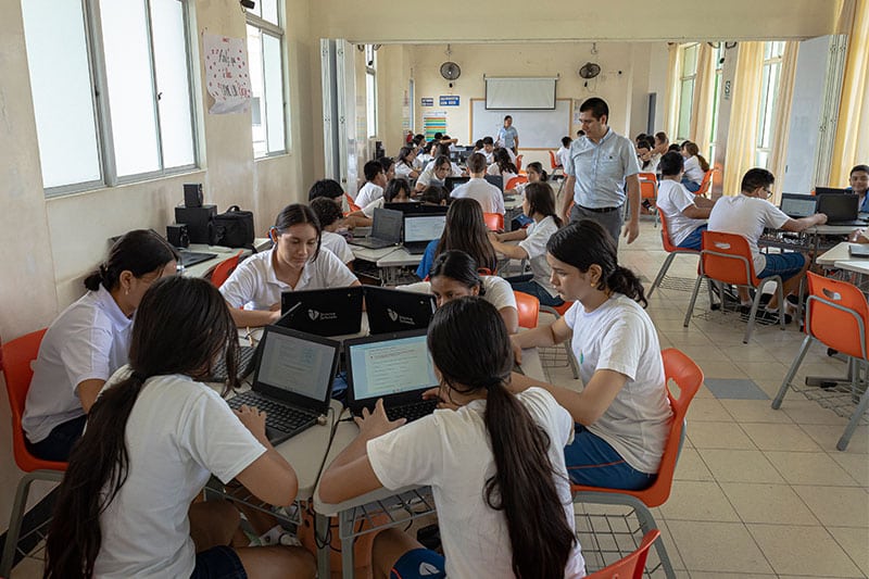 Students in a classroom work collaboratively on laptops in small groups, while teachers observe and assist.