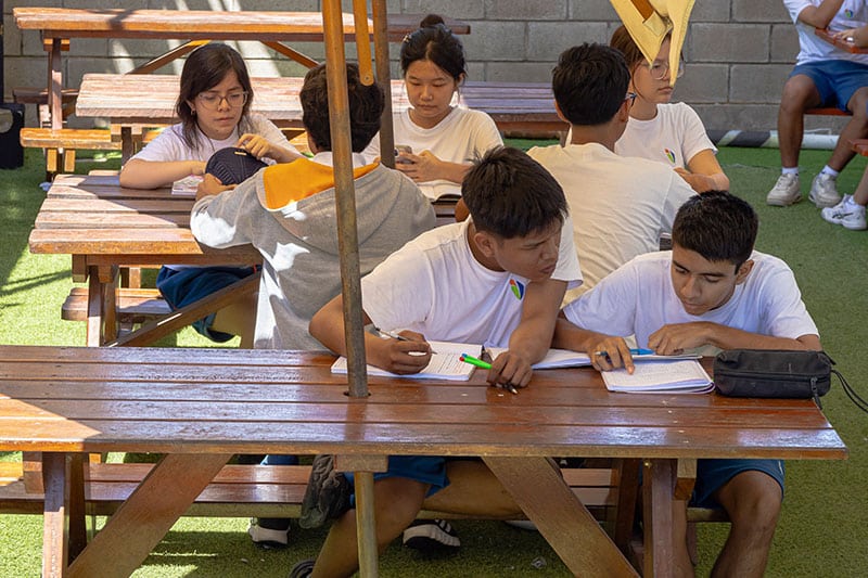 Groups of students in white shirts study and collaborate at wooden picnic tables under umbrellas in an outdoor school area with artificial grass.